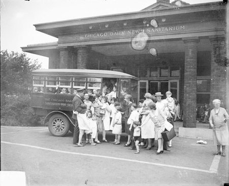 Children and adults standing in front of a crowded bus in front of the Chicago Daily News Sanitarium; a man wearing a uniform is standing amid the crowd, with his arm around a child's shoulder - Chicago Daily News, Inc.