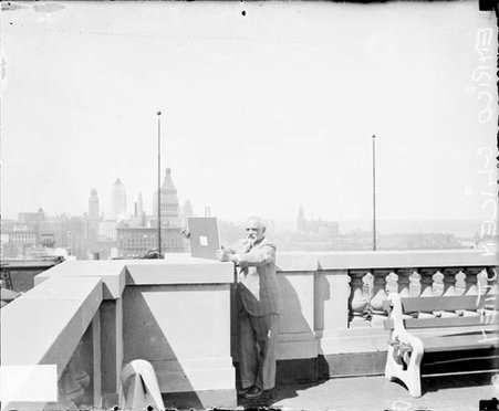 Enrico Glicenstien holding a flat, rectangular object, standing on the large balcony of a building - Chicago Daily News, Inc.