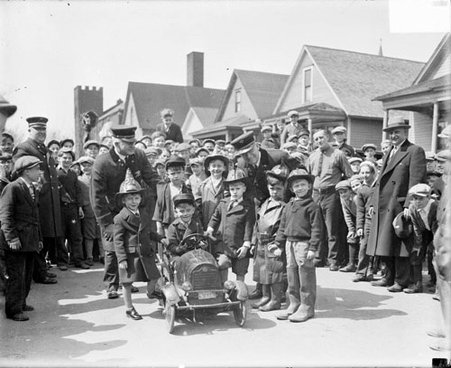 Young boy sitting in a miniature fire truck on a street, surrounded by a group of boys and some men - Chicago Daily News, Inc.