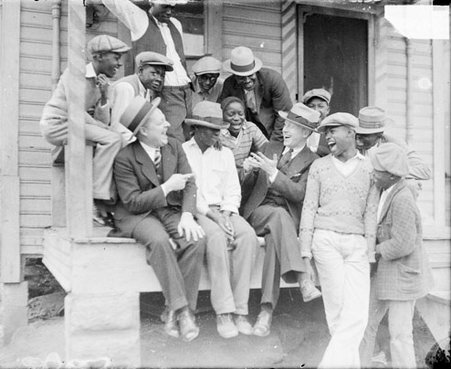 WMAQ radio actors, Charles J. Correll and Freeman Gosden of Amos 'n Andy, sitting and laughing with a group of African American men and boys on a porch in front of a house, light exposure - Chicago Daily News, Inc.