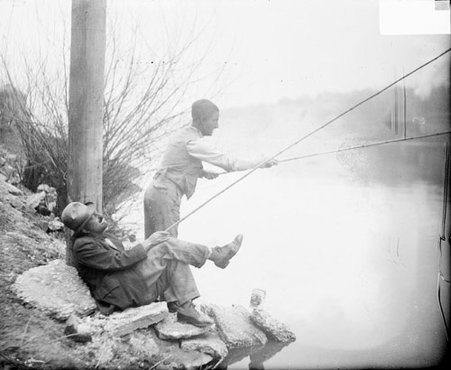 WMAQ radio actors, Charles J. Correll and Freeman Gosden of Amos 'n Andy, wearing blackface and costumes, standing and sitting while fishing in a body of water, light exposure - Chicago Daily News, Inc.