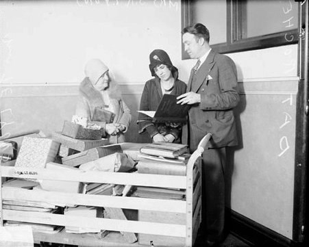 Mrs. T. R. Lyons, Mrs. S. R. Lyons, and Sergeant Pat Roche standing behind a cart of books and boxes in a room - Chicago Daily News, Inc.