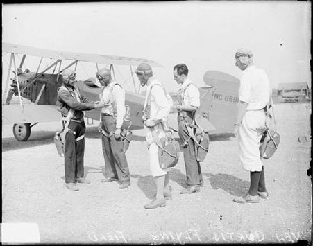 Five men wearing flight gear and parachutes standing in front of an airplane on an airfield - Chicago Daily News, Inc.
