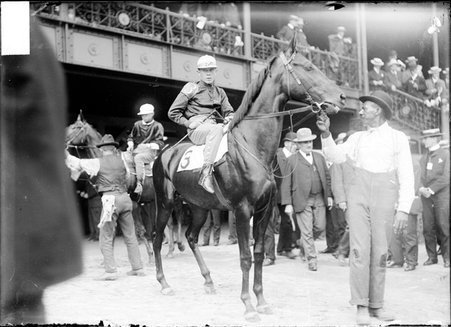 Racehorse, Tryon, with jockey mounted and African American handler holding bridle, standing on paddock by grandstand - Chicago Daily News, Inc.