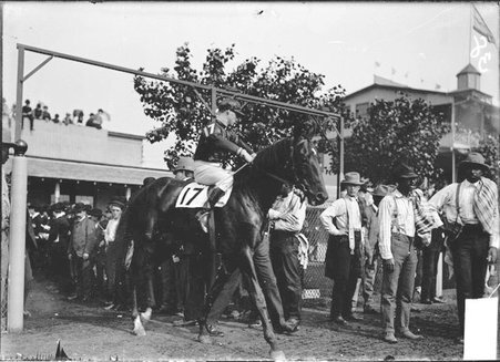 Racehorse, Jordan, with mounted jockey, walking through a racetrack paddock gate past horsemen and handlers - Chicago Daily News, Inc.