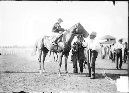 Racehorse, Filipe Lugos, standing on a racetrack, jockey sitting in saddle, handlers assisting - Chicago Daily News, Inc.