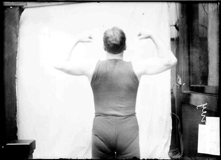 Wrestler, Rooney, flexing muscles, facing away from the camera, standing against a plain backdrop - Chicago Daily News, Inc.
