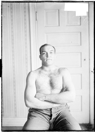 Pugilist, James J. Jeffries, facing camera, arms folded over lower chest, sitting in a room, far shot - Chicago Daily News, Inc.