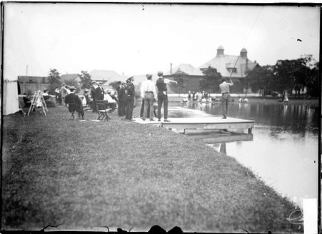 Fly fishing scene, fisherman casting from a pier, spectators watching from the lake bank - Chicago Daily News, Inc.