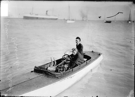 Driver and passenger riding in the motorboat Crusader on Lake Michigan, moored sailboats and a steamship are visible in the background - Chicago Daily News, Inc.