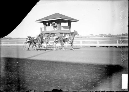 Racehorse, Danube, and driver leading two other racehorses and drivers as they are passing brick tower - Chicago Daily News, Inc.