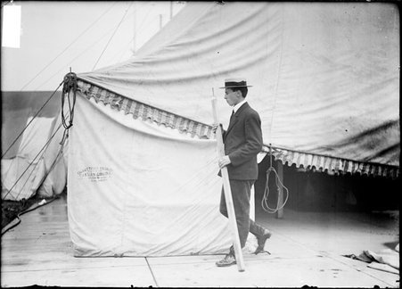 A. Roy Knabenshue, airship builder, carrying a pole and passing a tent which housed an airship - Chicago Daily News, Inc.