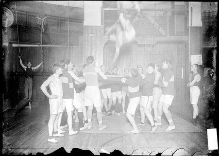 Athletes holding on to the edges of a piece of material and throwing another young man into the air in a gymnasium at the YMCA - Chicago Daily News, Inc.