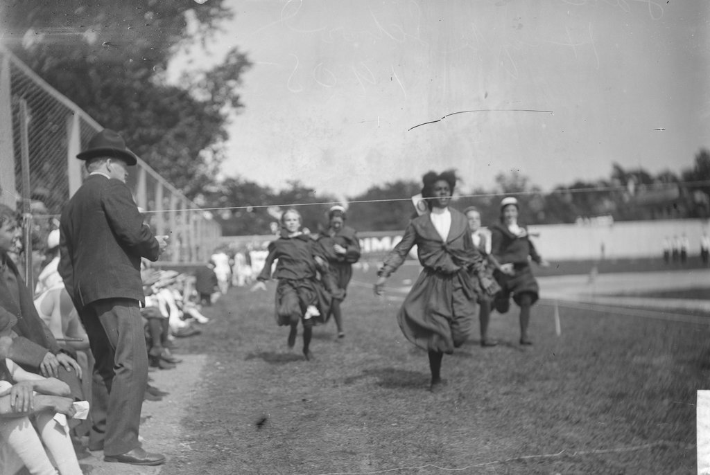 African American girl crossing the finish line ahead of four Caucasian girls during a running race on the grass at an athletic field - Chicago Daily News, Inc.