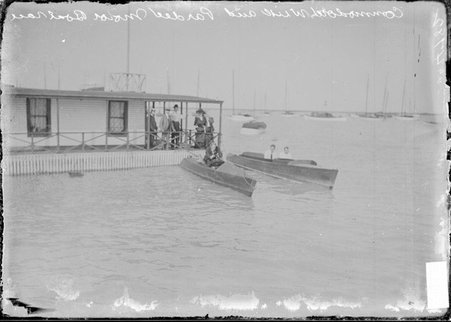 Commodore Theodore Weise and F. J. Pardee, each sitting at the wheel of a motorboat preparing for a race near a boathouse in a harbor - Chicago Daily News, Inc.