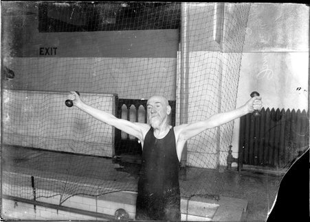 Swimmer, James M. Angus, standing with his arms stretched out to the sides holding small weights in the corner of a pool in a gymnasium - Chicago Daily News, Inc.