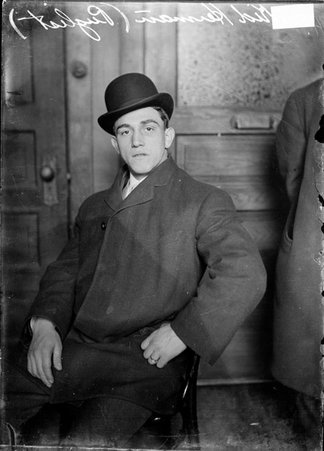 Boxer Kid Herman, wearing an overcoat and a hat, sitting in an office, the shoulder of a person in the background is partially visible - Chicago Daily News, Inc.