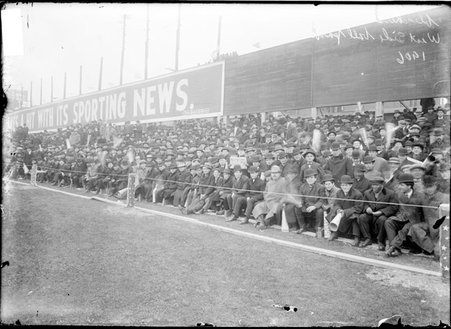 Spectators sitting in bleachers behind a fence at West Side Grounds, billboard above bleachers carries a message about the Tribune - Chicago Daily News, Inc.