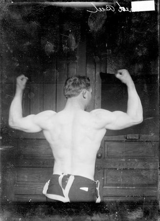 Wrestler, Fred Beell, standing with his back to the camera flexing his arm muscles with fists turned up to ceiling in a room - Chicago Daily News, Inc.