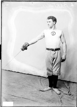 Fencer G. Barth, holding a foil and pointing it towards the ground, standing in front of a light-colored backdrop in a room - Chicago Daily News, Inc.
