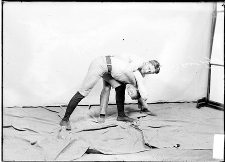 Wrestlers Henry Legel and Fred Richelsen, Legel holding Richelsen in a full-nelson, standing in front of a light-colored backdrop - Chicago Daily News, Inc.
