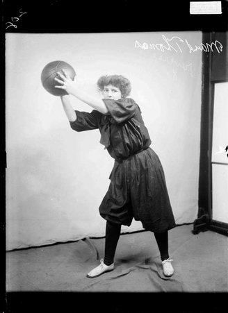 Basketball player, Maud Thomas, holding basketball at head level with both hands in front of a light colored backdrop in a room - Chicago Daily News, Inc.