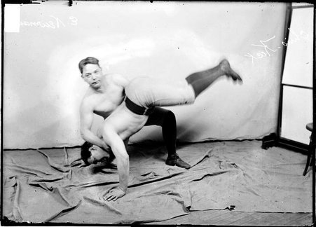 Wrestlers Newman and Keck, Newman is holding Keck belly-first over his knee by his waist and neck, kneeling in front of a backdrop - Chicago Daily News, Inc.