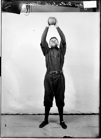Indoor baseball player, De LaSalle, Egan, holding an indoor baseball above his head with both hands, standing in front of a backdrop - Chicago Daily News, Inc.