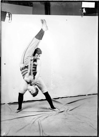 Wrestlers, Louis Bowman and W. J. Weir, Bowman holding Weir upside down, standing in front of a backdrop - Chicago Daily News, Inc.