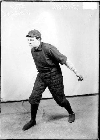 De LaSalle indoor baseball player J. McHugh, extending his left arm behind him, standing in front of a light-colored backdrop in a room - Chicago Daily News, Inc.