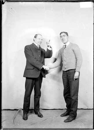 Basketball players, C. A. Torizalin and A. L. Mac Bear, shaking hands while one of them points in front of a backdrop in a room - Chicago Daily News, Inc.