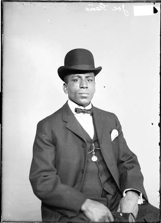 Boxer, Joe Gans, wearing a hat and sitting in a chair in front of a light colored backdrop in a room - Chicago Daily News, Inc.