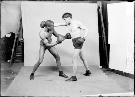 Boxer, Joe Gans, standing with legs spread apart, punching opponent in the stomach in front of a backdrop - Chicago Daily News, Inc.