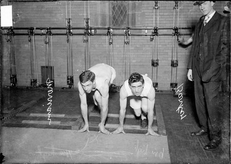 Runners, University of Chicago, Merriam and Barker, in starting positions for running while an official stands nearby in a gymnasium - Chicago Daily News, Inc.