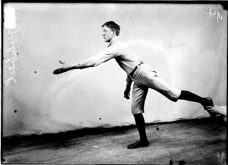 Indoor baseball player, R. Holnbeck, leaning forward with his left arm extended in front of him, standing on one leg in front of a light-colored backdrop - Chicago Daily News, Inc.