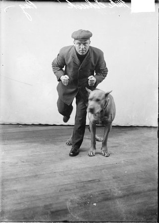 Pugilist Mike Schieck, wearing street clothes and leaning forward, standing on one leg in front of a backdrop, dark exposure - Chicago Daily News, Inc.