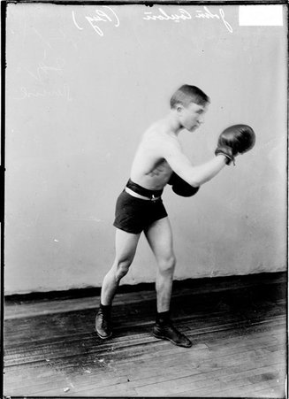 Pugilist John Coulon, standing in profile in a boxing stance in front of a light-colored backdrop in a room - Chicago Daily News, Inc.