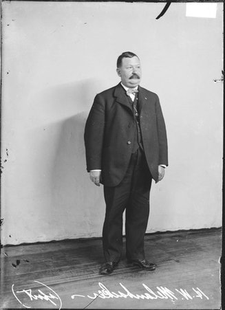 H. W. Melanbacker wearing a suit, standing in front of a light-colored backdrop in a room - Chicago Daily News, Inc.