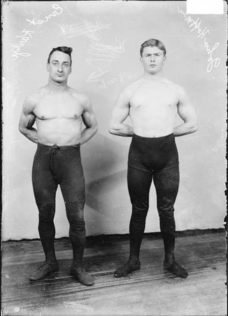 Wrestlers Ernest Rartye and John Hoffman, standing in front of a light-colored backdrop in a room - Chicago Daily News, Inc.