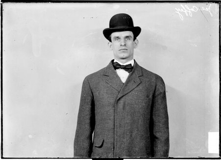 Pugilist Joe Coffey, wearing a hat and suit, standing in front of a light-colored backdrop in a room - Chicago Daily News, Inc.