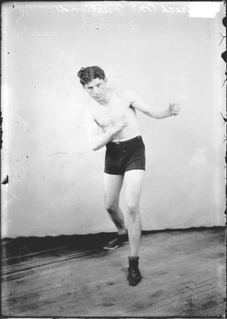 Pugilist Patrick McFarland, facing the camera, standing in a boxing stance in front of a backdrop in a room, dark exposure - Chicago Daily News, Inc.