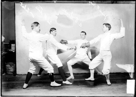 Fencers Eckart, Stone, Sauer, and Knox, holding foils at one another, standing in fencing stances in front of a backdrop - Chicago Daily News, Inc.