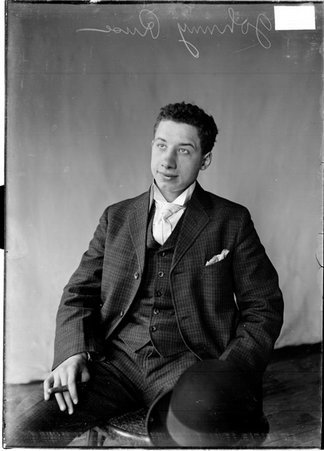 Johnny Russ wearing a suit and holding a cigar, sitting in front of a light-colored backdrop in a room - Chicago Daily News, Inc.