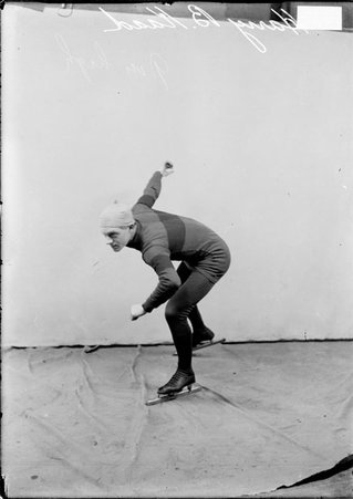 Skater Harry B. Raad, wearing ice skates and skating clothing, crouching in a ready position in front of a backdrop in a room - Chicago Daily News, Inc.