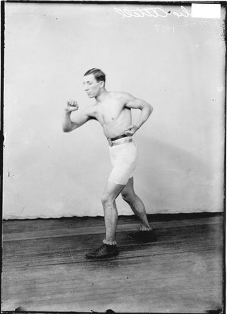 Pugilist Abe Attell, standing in a boxing stance in front of a light-colored backdrop in a room - Chicago Daily News, Inc.