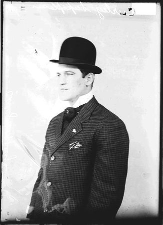 Curley Supples, wearing a suit and hat and facing to the left of the camera, sitting in front of a light-colored backdrop in a room - Chicago Daily News, Inc.