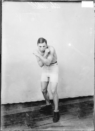 Pugilist Abe Attell, crossing his arms in front of his chest, standing in a boxing stance in front of a backdrop in a room - Chicago Daily News, Inc.