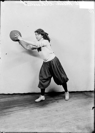 Spalding girls' basketball captain Meta Schmidt, holding a basketball, extended in front of her, standing in front of a backdrop - Chicago Daily News, Inc.