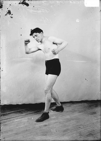 Pugilist Patrick McFarland, drawing his left arm back, standing in a boxing stance in front of a backdrop in a room - Chicago Daily News, Inc.