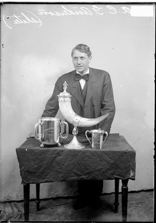 Skater Adolph C. G. Anderson, wearing a suit, standing behind a table in front of a backdrop, light exposure - Chicago Daily News, Inc.